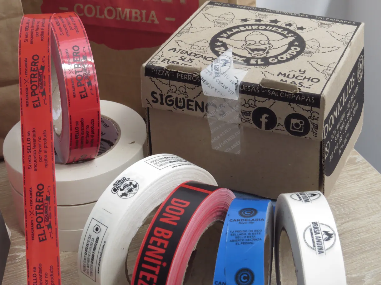Caja de comida a domicilio precintada con la cinta de seguridad, junto a los rollos de varias marcas