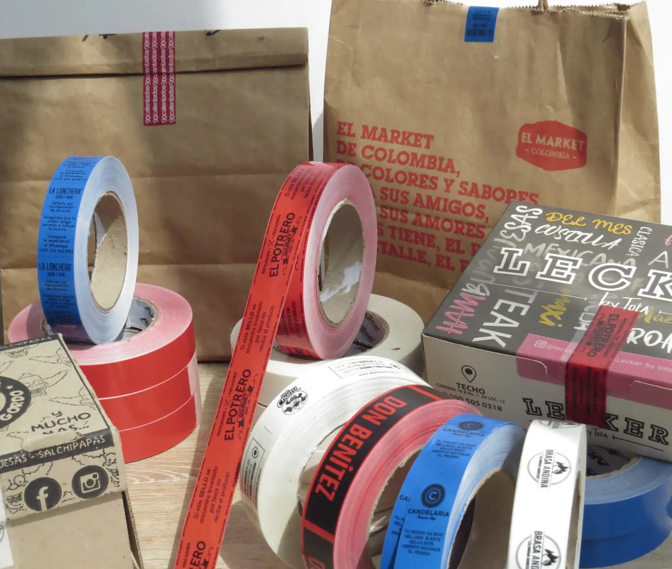 Bolsas de papel de reparto a domicilio precintadas, junto a rollos de cinta de seguridad personalizada en varios colores